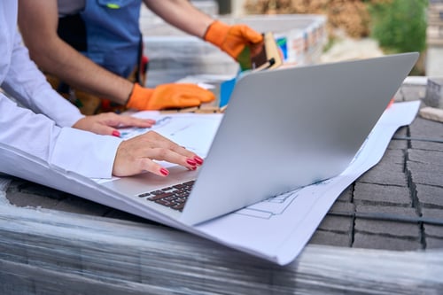 Is Roofing Business Profitable? 5 Person using a laptop on roofing plans while discussing costs and the question is roofing business profitable.